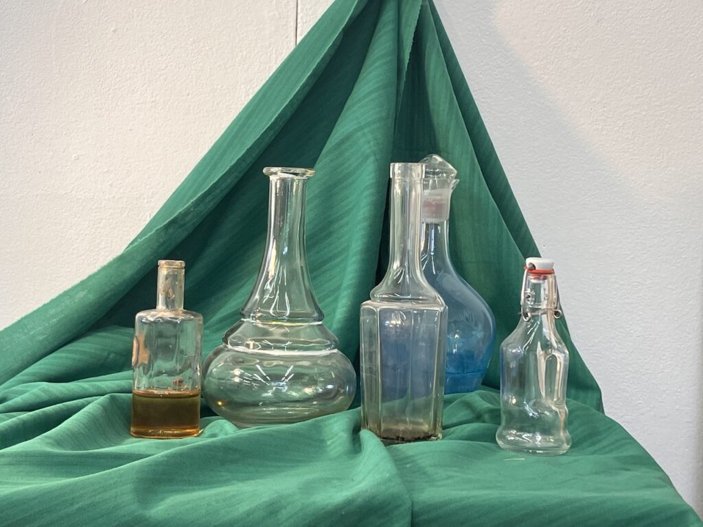 Still life for a colour pencil drawing, with a set of glass bottles against a green background, with lots of reflections